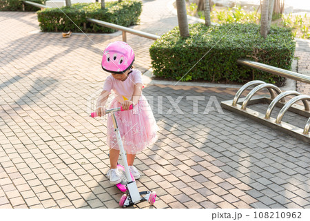 Child riding scooter. Happy Asian little kid girl wear safe helmet playing pink kick board on road in park outdoors on summer day, Active children games outside, Kids sport healthy lifestyle concept Child riding scooter. Happy Asian little kid girl wear safe helmet playing pink kick board on road in park outdoors on summer day, Active children games outside, Kids sport healthy lifestyle concept 108210962