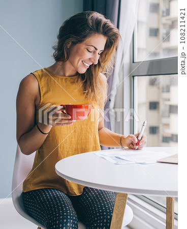 Laughing female sitting at table with notebook and cup filling documents Laughing female sitting at table with notebook and cup filling documents 108211421