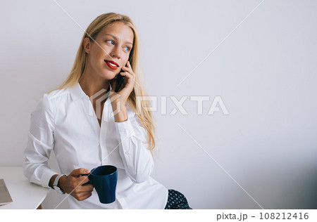Focused female sitting at table talking on phone and working on laptop 108212416