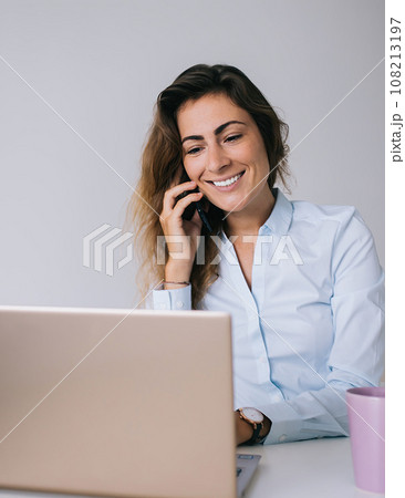 Smiling young woman talking on smartphone and typing on laptop 108213197