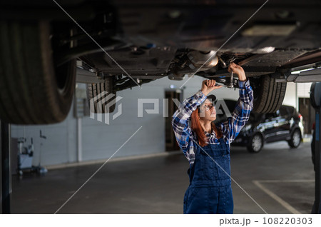 Female mechanic unscrew the nuts on the bottom of the car that is on the lift. A girl at a man's work. Female mechanic unscrew the nuts on the bottom of the car that is on the lift. A girl at a man's work. 108220303
