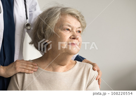 Older woman looking into distance, therapist touch shoulders supporting patient 108220554