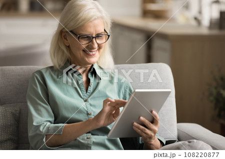 Positive elderly woman using digital gadget on home sofa Positive elderly woman using digital gadget on home sofa 108220877