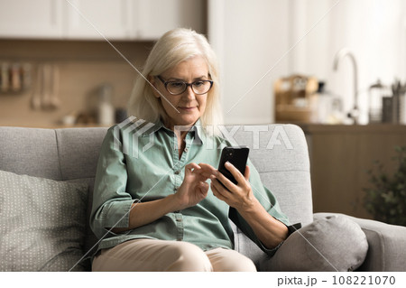 Serious elderly blonde woman in eyewear typing on cellphone 108221070