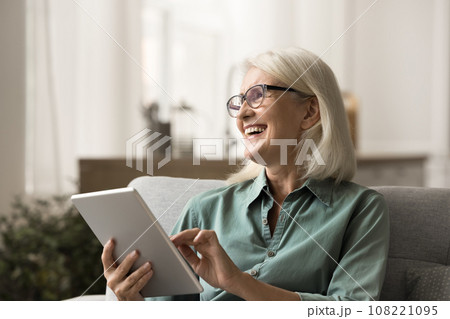 Cheerful elderly woman resting on home sofa with tablet 108221095