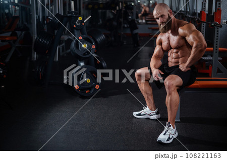 A muscular bald man in shorts is resting on a bench after a workout. Bodybuilder showing off his shape in the gym. A muscular bald man in shorts is resting on a bench after a workout. Bodybuilder showing off his shape in the gym. 108221163