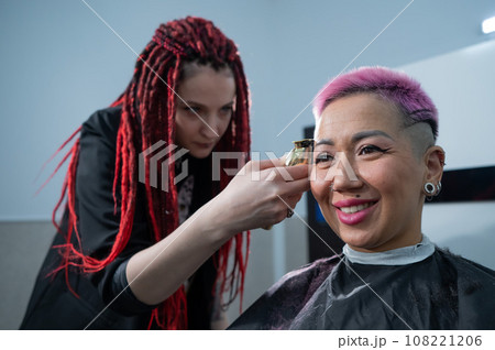 The hairdresser shaves the temple of a female client. Rear view of a woman with short pink hair in a barbershop. 108221206