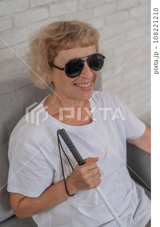 An elderly blind woman wearing sunglasses and with a tactile cane sits on the sofa. An elderly blind woman wearing sunglasses and with a tactile cane sits on the sofa. 108221210