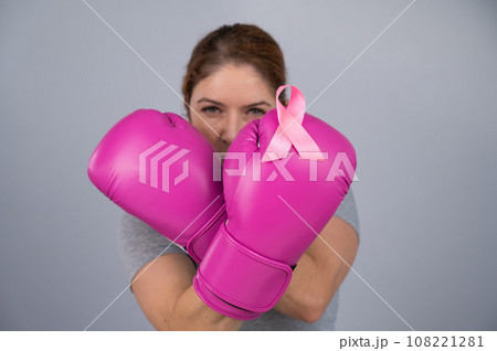 Caucasian woman in pink boxing gloves with a pink ribbon on her chest on a gray background. Fight against breast cancer.  108221281