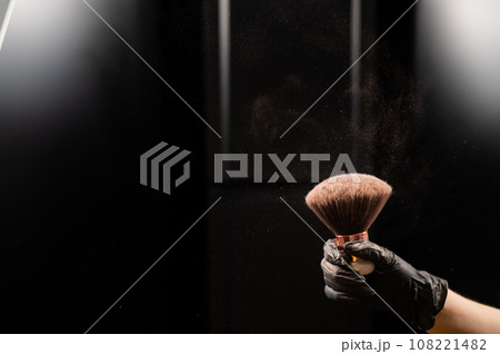 A woman shakes a large powder brush with clouds of dust on a black background. Treatment after instant tanning 108221482
