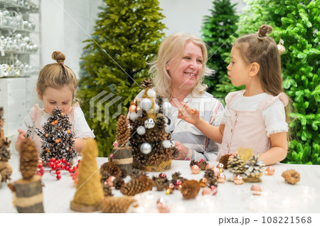 Elderly caucasian woman making pine cones decoration for christmas with two granddaughters Elderly caucasian woman making pine cones decoration for christmas with two granddaughters 108221568