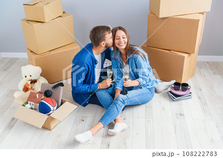 Couple taking a break on moving day, sitting on floor in new apartment celebrating with champagne surrounded by cardboard boxes. Couple taking a break on moving day, sitting on floor in new apartment celebrating with champagne surrounded by cardboard boxes. 108222783