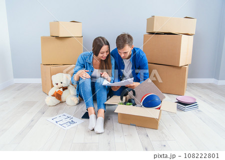 Couple have a discussion about design of their new home sitting on the wooden floor between many cardboard boxes. Beautiful young stylish couple in love planning home design for their new bought flat. Couple have a discussion about design of their new home sitting on the wooden floor between many cardboard boxes. Beautiful young stylish couple in love planning home design for their new bought flat. 108222801