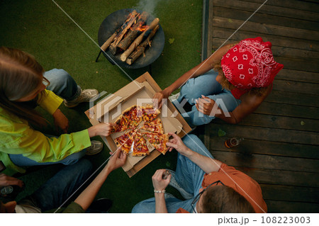 Overhead view on group of friends eating pizza nearby burning campfire 108223003