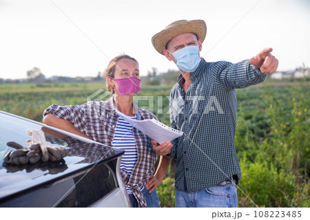 Farmer couple in protective masks discussing papers near car 108223485