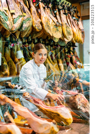 Young salesgirl in white uniform cutting jerky ham for sale in butcher shop 108223515