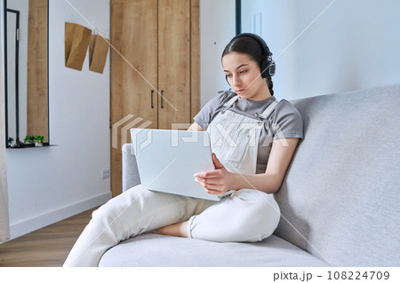 Teen girl in headphones at home on couch using laptop 108224709