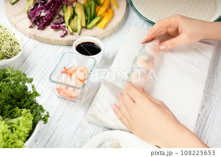 Woman making rice paper roll at white wooden table, closeup Woman making rice paper roll at white wooden table, closeup 108225653