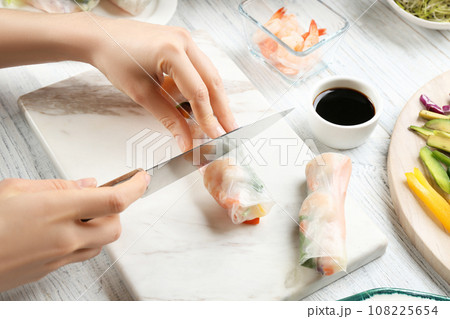 Woman cutting rice paper roll at white wooden table, closeup Woman cutting rice paper roll at white wooden table, closeup 108225654