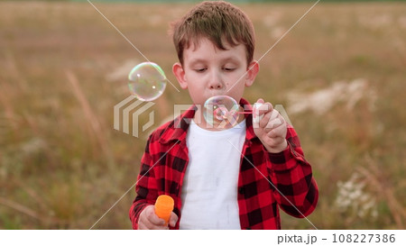 Happy boy blows soap bubbles from bottle in autumn field of crops closeup Happy boy blows soap bubbles from bottle in autumn field of crops closeup 108227386