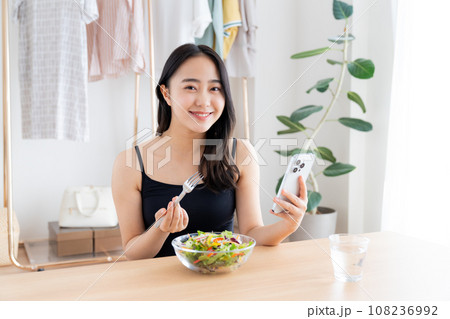 スマホを見ながらサラダを食べる若い女性 スマホを見ながらサラダを食べる若い女性 108236992
