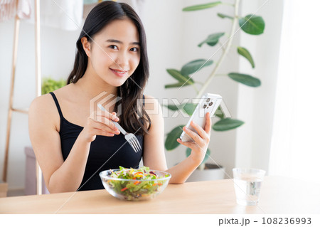 スマホを見ながらサラダを食べる若い女性 スマホを見ながらサラダを食べる若い女性 108236993