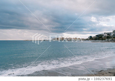 Sea waves roll on the sandy beach against the backdrop of a stormy sky and high-rise buildings on the shore 108237691