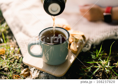Coffee pours from a thermos into a mug standing on a wooden plank on the lawn Coffee pours from a thermos into a mug standing on a wooden plank on the lawn 108237820