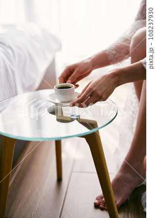 Bride takes a cup of coffee from a glass table while sitting in an armchair. Cropped. Faceless Bride takes a cup of coffee from a glass table while sitting in an armchair. Cropped. Faceless 108237930