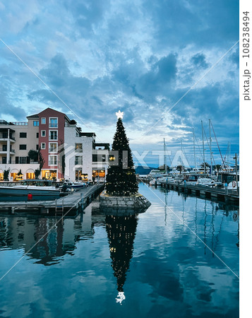 Christmas tree decorated with luminous garlands stands on the pier against the backdrop of houses and moored yachts 108238494
