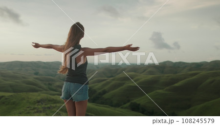 Woman enjoy sunset landscape, raises hands against green hills valley. Long hair brunette girl looking on beautiful nature scene. Freedom, happiness lifestyle. Travel, tourism, holiday. Woman enjoy sunset landscape, raises hands against green hills valley. Long hair brunette girl looking on beautiful nature scene. Freedom, happiness lifestyle. Travel, tourism, holiday. 108245579