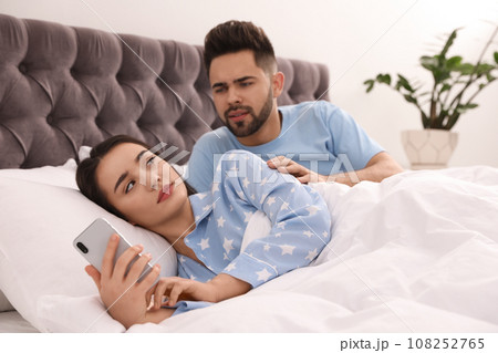 Young woman ignoring her distrustful boyfriend while using smartphone in bed at home 108252765