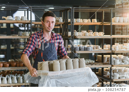 Caucasian male potter holding tray with ceramic pots in the pottery. Caucasian male potter holding tray with ceramic pots in the pottery. 108254787