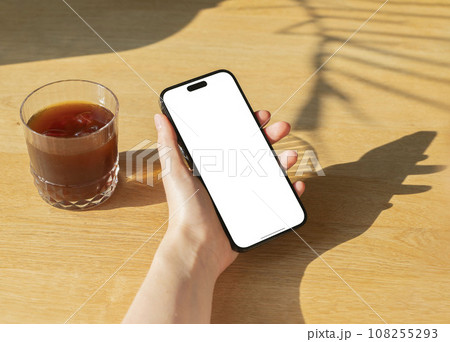 Berlin, Germany October 20 2023 Hand holding mobile phone screen mock up on cafeteria table with cold coffee glass Berlin, Germany October 20 2023 Hand holding mobile phone screen mock up on cafeteria table with cold coffee glass 108255293
