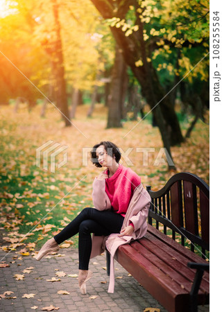 Young beautiful ballerina in fair coat and pointe shoes sit on the bench, rest outdoors. Young beautiful ballerina in fair coat and pointe shoes sit on the bench, rest outdoors. 108255584