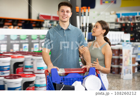 Spouses shopper stand with trolley filled with paint goods for repair and coloring in store sales hall. 108260212