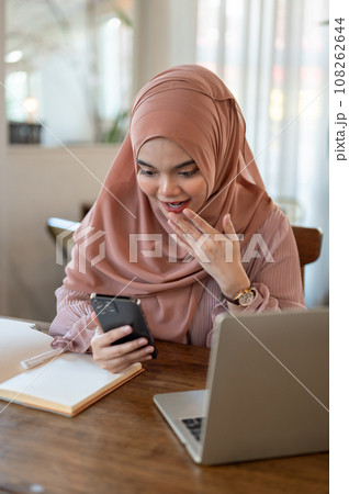 An excited Asian-Muslim woman is looking at her smartphone screen with a surprising face. 108262644