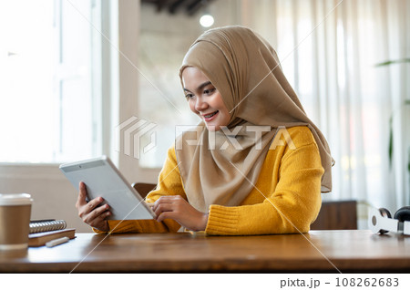 An attractive and happy Asian-Muslim woman is using a digital tablet while relaxing in a coffee shop 108262683