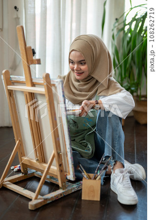 Happy Asian-Muslim female artist is sitting on the floor, enjoying creating her artwork on a canvas 108262719