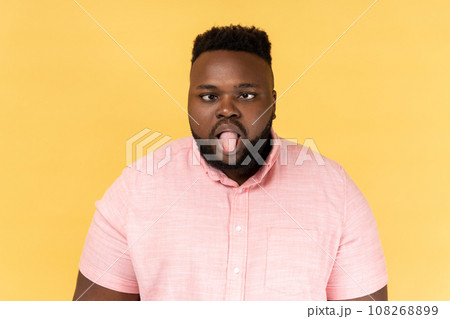 Portrait of disobedient crazy funny man wearing pink shirt showing tongue out and crossed his eyes displeased, behaving unruly naughty. Indoor studio shot isolated on yellow background. Portrait of disobedient crazy funny man wearing pink shirt showing tongue out and crossed his eyes displeased, behaving unruly naughty. Indoor studio shot isolated on yellow background. 108268899