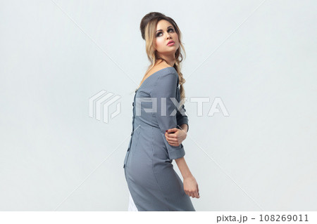 Beautiful blonde woman standing touching her hand looking away, fashion model with perfect body and attractive appearance, wearing grey elegant dress. Indoor studio shot isolated on gray background. 108269011