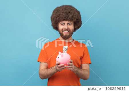 Portrait of smiling man with Afro hairstyle wearing orange T-shirt holding piggy bank with dollar banknote in hands, being happy of savings. Indoor studio shot isolated on blue background. Portrait of smiling man with Afro hairstyle wearing orange T-shirt holding piggy bank with dollar banknote in hands, being happy of savings. Indoor studio shot isolated on blue background. 108269013