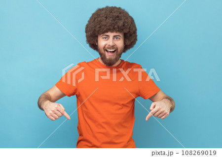 Portrait of excited satisfied man with Afro hairstyle wearing orange T-shirt pointing down with fore fingers, looking at camera with toothy smile. Indoor studio shot isolated on blue background. 108269019