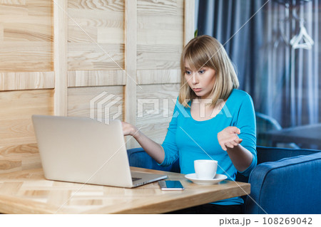 Portrait of confused uncertain young woman with blonde hair in blue shirt sitting at table and working on laptop, shrugging shoulders, looks doubtful and puzzled. Indoor shot, cafe background. Portrait of confused uncertain young woman with blonde hair in blue shirt sitting at table and working on laptop, shrugging shoulders, looks doubtful and puzzled. Indoor shot, cafe background. 108269042