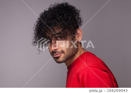 Portrait of attractive handsome young adult bearded man with curly hair, wearing red jumper, looking at camera, showing his hairdo. Indoor studio shot isolated on gray background. Portrait of attractive handsome young adult bearded man with curly hair, wearing red jumper, looking at camera, showing his hairdo. Indoor studio shot isolated on gray background. 108269043