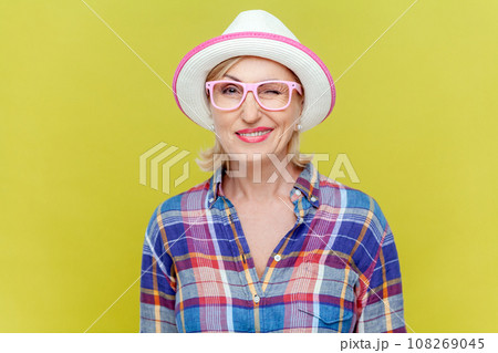 Cheerful playful mature woman in shirt, hat and eyeglasses, being in good mood, smiling broadly and winking at camera, feelings, attitude and reaction. Indoor studio shot isolated on yellow background Cheerful playful mature woman in shirt, hat and eyeglasses, being in good mood, smiling broadly and winking at camera, feelings, attitude and reaction. Indoor studio shot isolated on yellow background 108269045