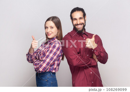 Satisfied delighted woman and man standing together showing thumbs up, like gesture, positive feedback, looking at camera with happy expression. Indoor studio shot isolated on gray background. Satisfied delighted woman and man standing together showing thumbs up, like gesture, positive feedback, looking at camera with happy expression. Indoor studio shot isolated on gray background. 108269099