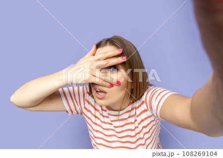 Portrait of spying woman in T-shirt taking selfie picture, point of view, looking at the camera through her finger, peeping, making front selfportrait. Indoor studio shot isolated on purple background Portrait of spying woman in T-shirt taking selfie picture, point of view, looking at the camera through her finger, peeping, making front selfportrait. Indoor studio shot isolated on purple background 108269104