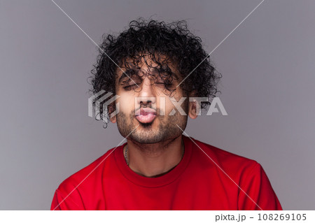 Portrait of romantic flirting handsome bearded man with curly hair, standing with closed eyes, keeps lips pout, sending kissing. wearing red jumper. Indoor studio shot isolated on gray background. Portrait of romantic flirting handsome bearded man with curly hair, standing with closed eyes, keeps lips pout, sending kissing. wearing red jumper. Indoor studio shot isolated on gray background. 108269105