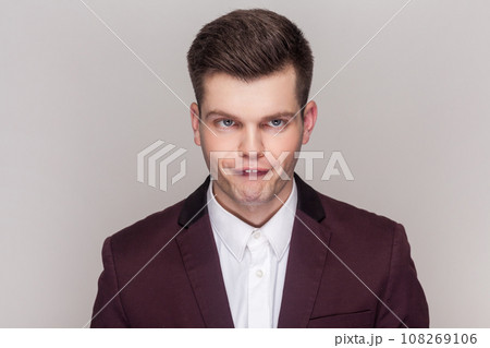 Portrait of crazy comic foolish handsome young man standing looking at camera with funny crossing eyes, wearing violet suit and white shirt. Indoor studio shot isolated on grey background. Portrait of crazy comic foolish handsome young man standing looking at camera with funny crossing eyes, wearing violet suit and white shirt. Indoor studio shot isolated on grey background. 108269106